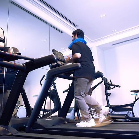 マカレム アイヤッド メッカ ホテル エクステリア 写真 The photo shows a person exercising on a treadmill in a modern gym or fitness room. The individual is wearing a blue neck support while jogging on the treadmill, which is set against a backdrop of large mirrors. There are also additional exercise equ