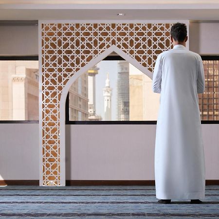 マカレム アイヤッド メッカ ホテル エクステリア 写真 The photo depicts a person standing with their back facing the viewer, wearing a traditional white garment. They are in a spacious interior setting that features an ornate archway with intricate patterns, typical of Islamic architecture. Through the 