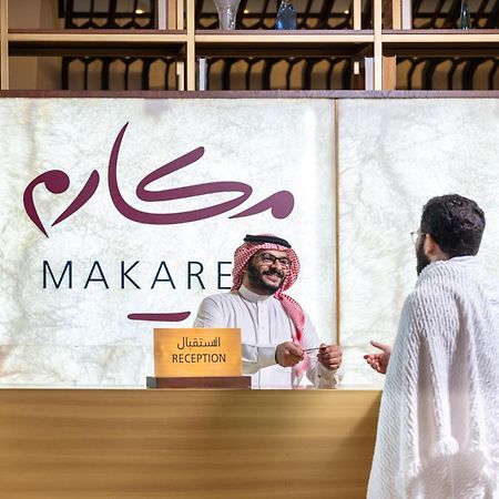 マカレム アイヤッド メッカ ホテル エクステリア 写真 The photo depicts a reception area in a hotel or establishment named "Makarem." In the image, a receptionist, dressed in traditional attire, is smiling and interacting with a guest who is wearing a white robe, commonly known as a thobe or ihram. The 
