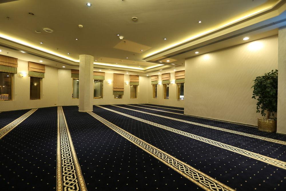 マカレム アイヤッド メッカ ホテル エクステリア 写真 The photo shows a spacious indoor area with a plush blue carpet featuring a decorative pattern. The room is well-lit, with soft lighting along the ceiling. There are several windows, allowing natural light to enter, and a few plants are positioned ar