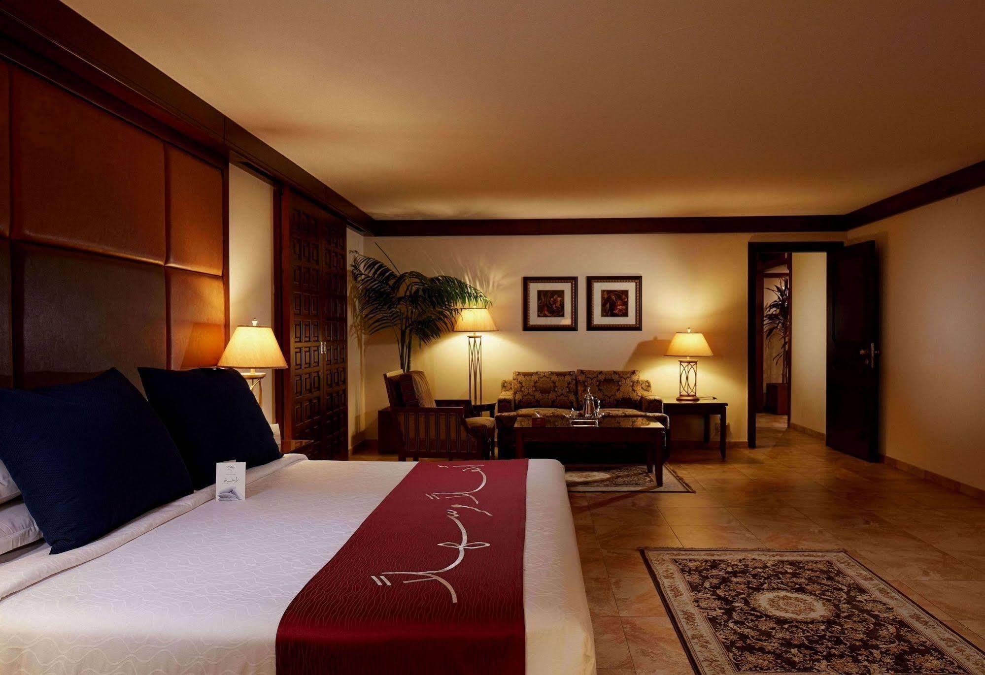 マカレム アイヤッド メッカ ホテル エクステリア 写真 The photo depicts a cozy hotel room with a warm and inviting atmosphere. It features a large bed with dark blue pillows and a maroon throw blanket. On either side of the bed, there are lamps providing soft lighting. In the background, a small seating