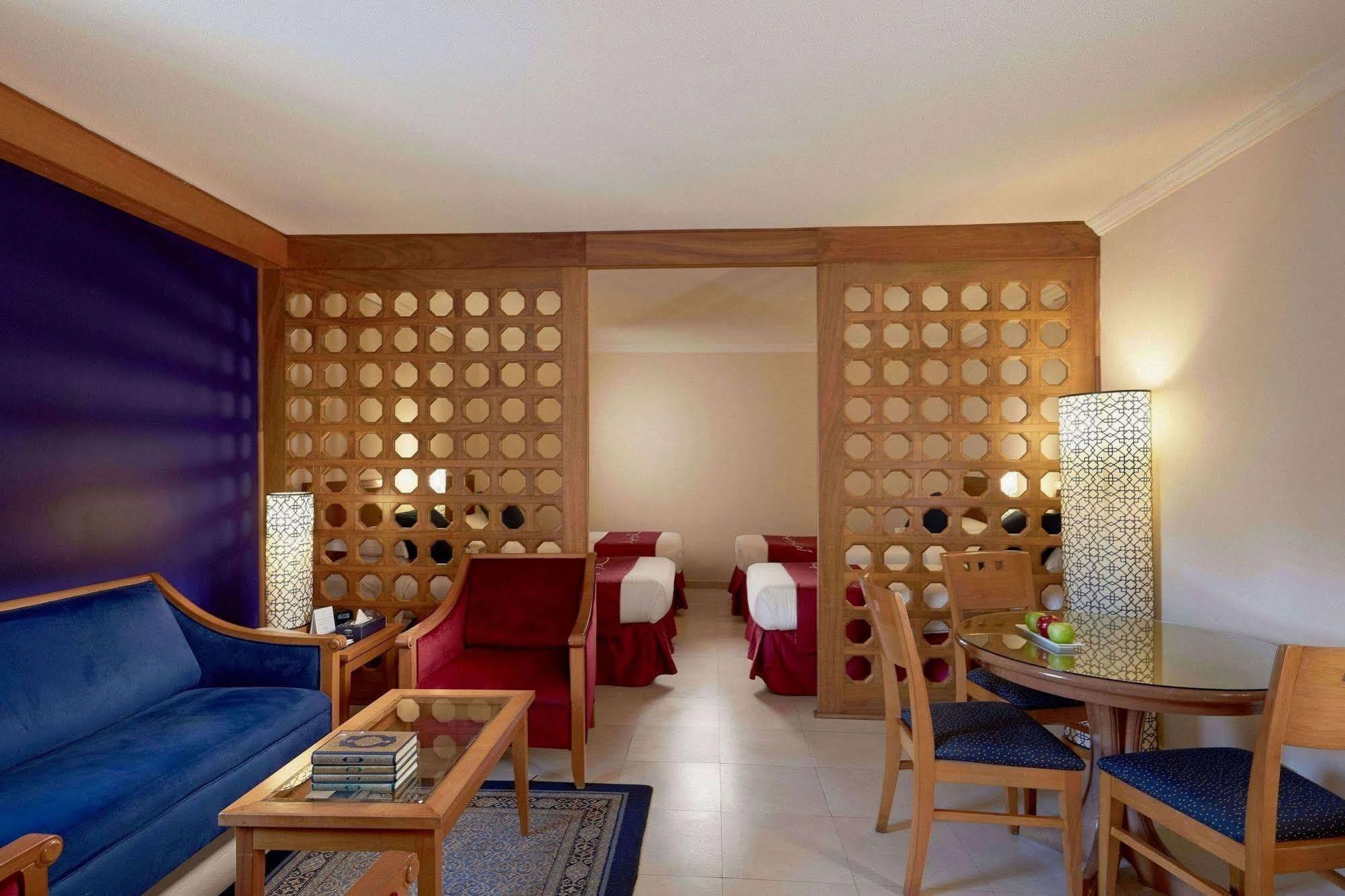マカレム アイヤッド メッカ ホテル エクステリア 写真 The photo shows a well-furnished hotel room or suite. On the left side, there is a seating area featuring a blue sofa and a small coffee table, as well as a couple of decorative items. The room is separated by a decorative wooden partition with circu