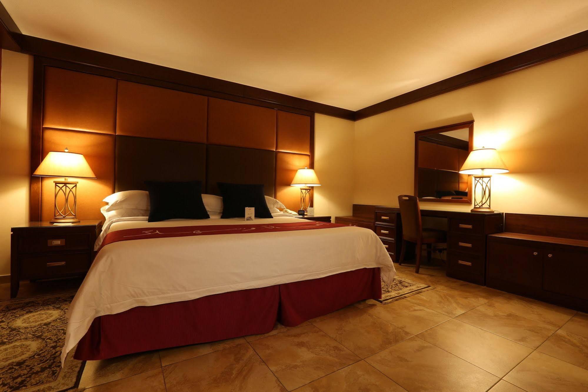 マカレム アイヤッド メッカ ホテル エクステリア 写真 The photo shows a well-appointed hotel room with a cozy and inviting atmosphere. It features a large bed with a white comforter and dark pillows, positioned against a wooden headboard. There are two bedside lamps providing soft lighting on either sid