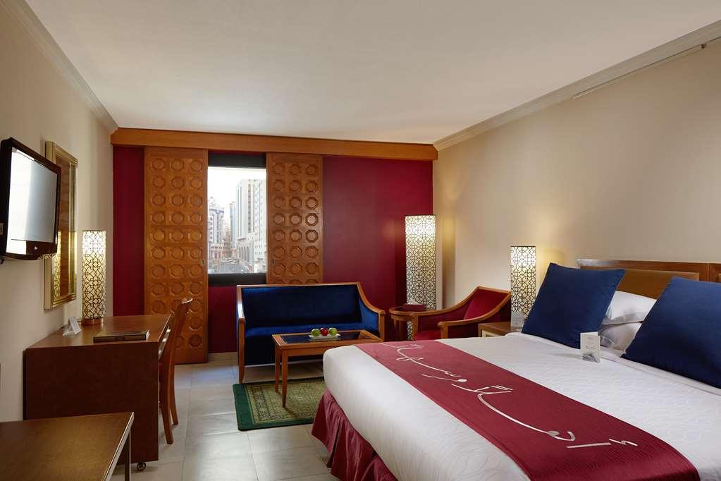 マカレム アイヤッド メッカ ホテル 部屋 写真 The photo showcases a hotel room featuring a modern design. In the center, there's a large bed with white linens and a decorative red runner. On either side of the bed, there are wooden nightstands with lamps. A blue sitting area is visible, composed