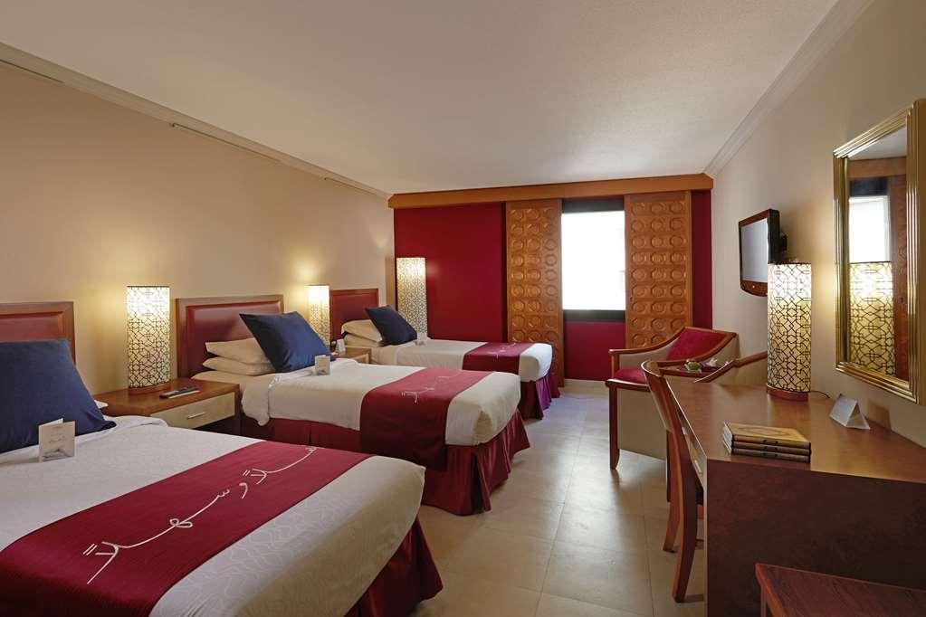 マカレム アイヤッド メッカ ホテル 部屋 写真 The photo shows a hotel room featuring three beds arranged with red and white bedding. The walls are painted in a neutral tone, and there are decorative lamps on nightstands beside each bed, providing ambient lighting. One of the walls has a window c