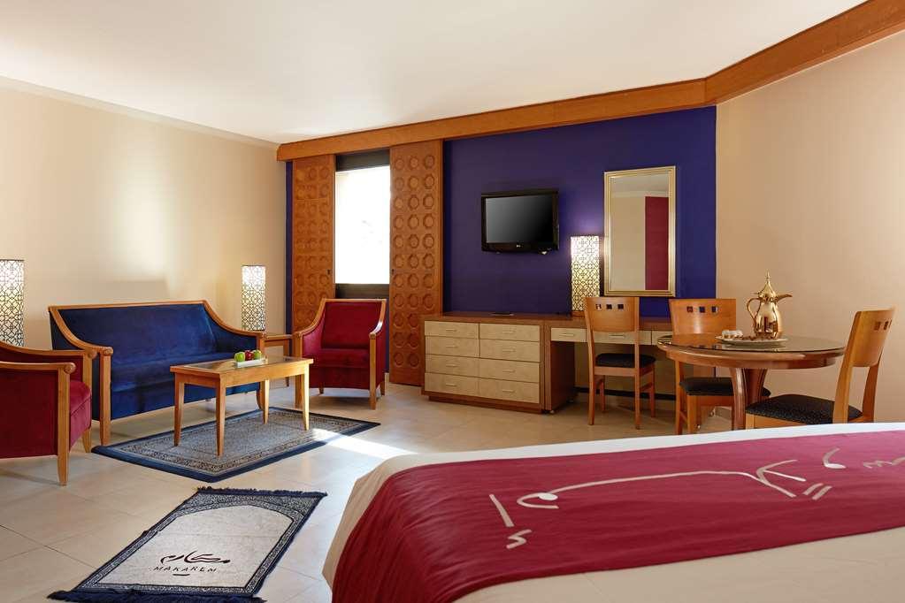 マカレム アイヤッド メッカ ホテル 部屋 写真 The image shows a hotel room with a modern, comfortable design. The room features a large bed with a red and white bedspread, a seating area that includes a blue sofa, two red chairs, and a wooden coffee table. There's a flat-screen TV mounted on the