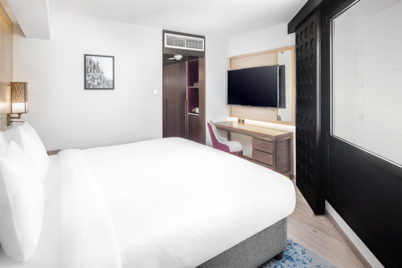 マカレム アイヤッド メッカ ホテル エクステリア 写真 The photo shows a modern hotel room featuring a large bed with white linens at the forefront. In the background, there is a wall-mounted television across from a desk with a chair. A wardrobe or cabinet is located to the side, and there's a decorativ