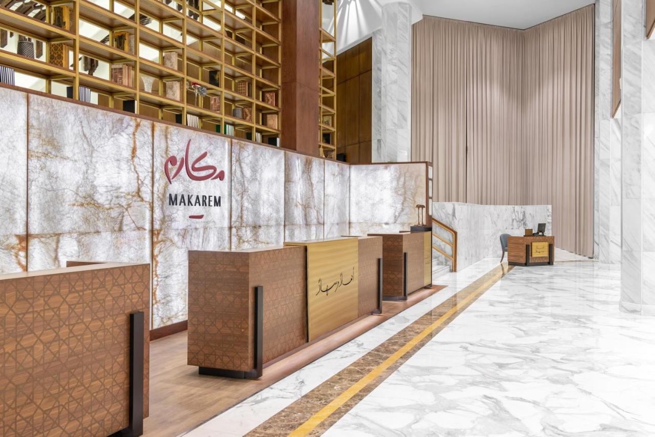 マカレム アイヤッド メッカ ホテル エクステリア 写真 The photo shows a modern hotel reception area with a sleek design. The reception desk is made of a combination of wood and stone, featuring a warm color palette. The backdrop is adorned with a marble finish, giving a luxurious feel. Above the desk, t