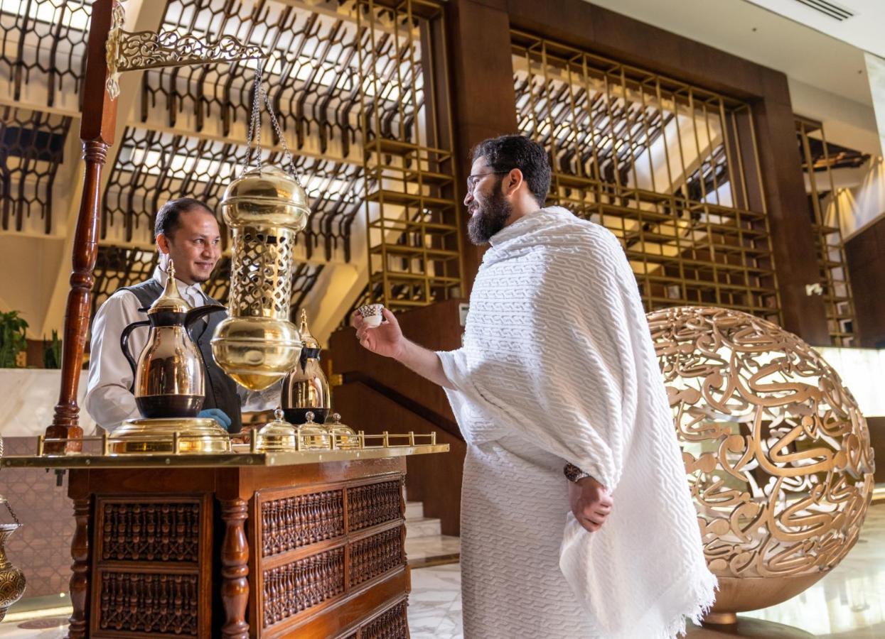 マカレム アイヤッド メッカ ホテル エクステリア 写真 The photo depicts a scene in a luxurious setting, likely a hotel or a fine dining establishment. In the foreground, a man wearing traditional attire, possibly a white garment, is engaging with a staff member. The staff member is behind a wooden count