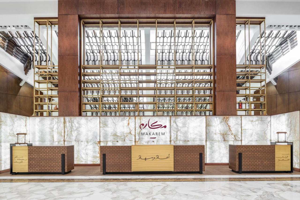 マカレム アイヤッド メッカ ホテル エクステリア 写真 The photo shows the interior of a hotel or a reception area, characterized by a modern and elegant design. In the foreground, there are multiple reception desks with a polished finish. Behind the desks, a striking feature is a wooden wall with a geom
