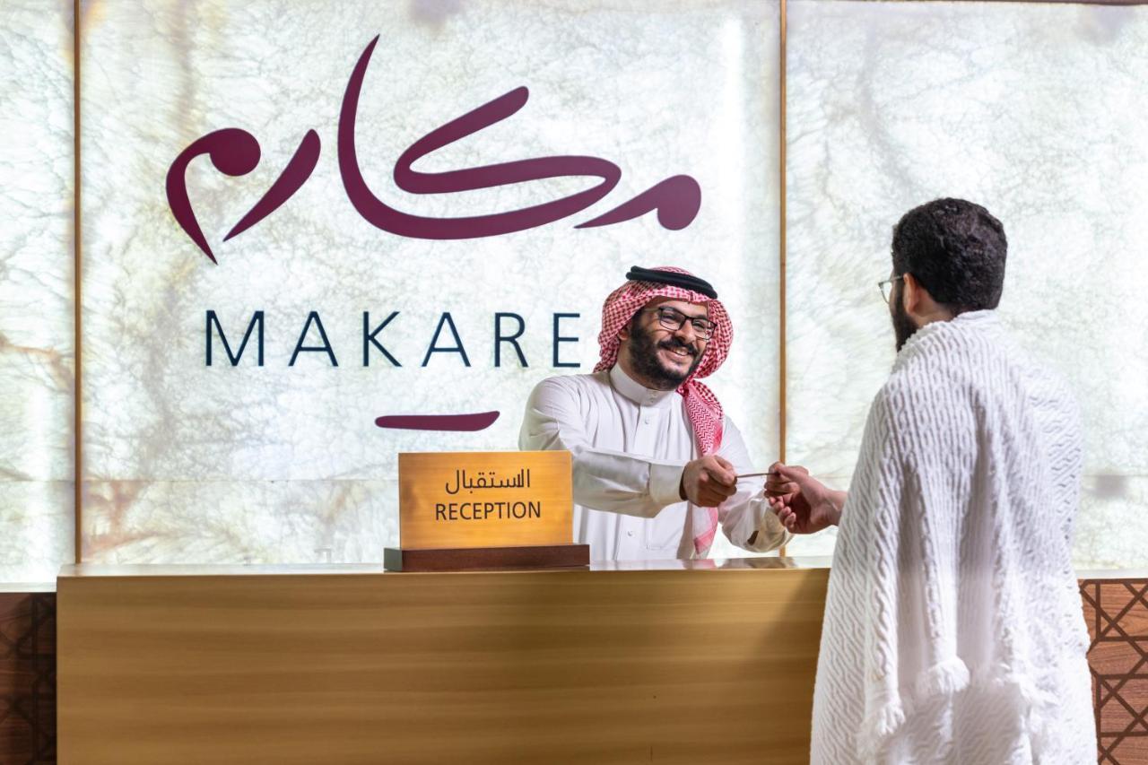 マカレム アイヤッド メッカ ホテル エクステリア 写真 The photo shows a reception area with a sign that reads "MAKARE" in both Arabic and English. In the foreground, a staff member wearing traditional attire is smiling and shaking hands with a visitor, who is clad in a white garment, typical for formal 
