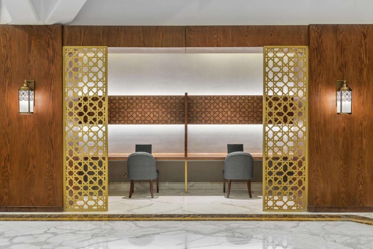 マカレム アイヤッド メッカ ホテル エクステリア 写真 The photo shows an elegantly designed space that features a reception or working area. There are two workstations with chairs positioned in front of them. The backdrop has a decorative panel with a geometric pattern. The area is framed by ornate gold