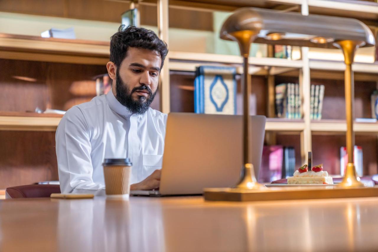 マカレム アイヤッド メッカ ホテル エクステリア 写真 The photo shows a man sitting at a desk in a library or a study area. He is wearing a white traditional garment and is focused on working on a laptop. In front of him, there is a cup of coffee and a small cake decorated with strawberries. The backgro