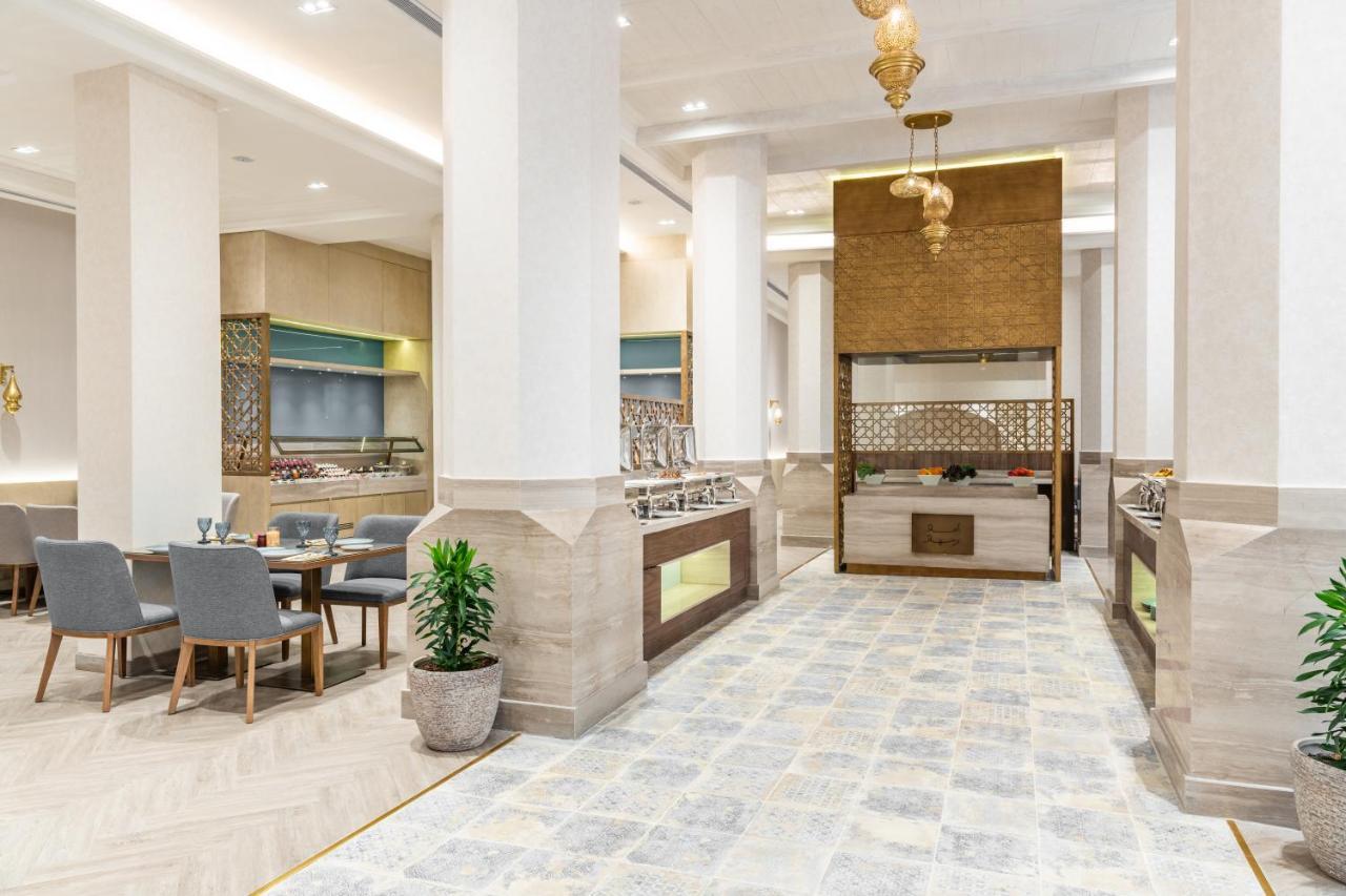 マカレム アイヤッド メッカ ホテル エクステリア 写真 The photo depicts a modern, elegant dining area, likely inside a restaurant or hotel. It features a spacious layout with tall ceilings and large columns. The flooring is a light-colored tiled design. 

To one side, there are several dining tables and