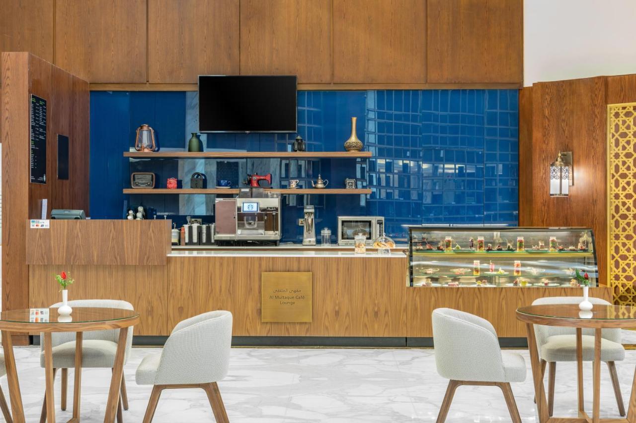 マカレム アイヤッド メッカ ホテル エクステリア 写真 The photo shows the interior of a café. The design features warm wood paneling and a striking blue tiled wall. In the foreground, there are several light-colored chairs and small tables arranged neatly. Behind the counter, there's a variety of coffee