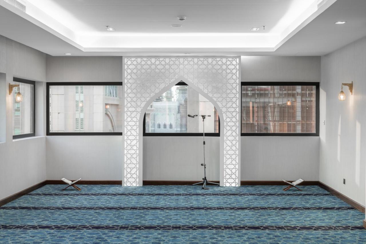 マカレム アイヤッド メッカ ホテル エクステリア 写真 The image shows a prayer room or small mosque interior. It features a white decorative wall with an archway that is typical in Islamic architecture. The floor is covered with a blue carpet, arranged with patterned lines, likely indicating prayer spot