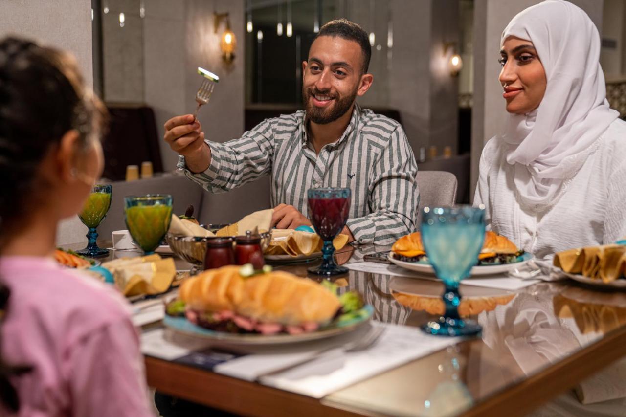 マカレム アイヤッド メッカ ホテル エクステリア 写真 The photo shows a dining scene with three people enjoying a meal together. A man is seated at the table, smiling and holding a utensil, possibly ready to serve or share food. He is dressed in a striped shirt. Across from him is a woman wearing a whit