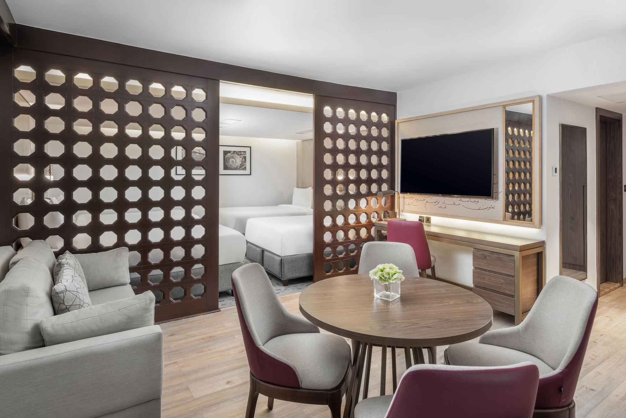 マカレム アイヤッド メッカ ホテル エクステリア 写真 The photo shows a modern hotel room or suite. It features a living area with a comfortable gray sofa and a round wooden dining table surrounded by upholstered chairs. A large flat-screen TV is mounted on the wall opposite the seating area. There's a 