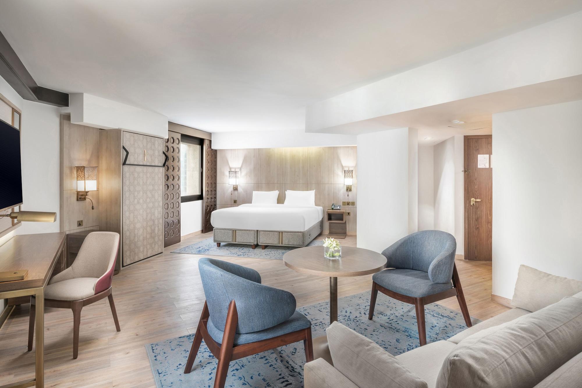 マカレム アイヤッド メッカ ホテル エクステリア 写真 The photo shows a modern hotel room with a spacious layout. It features a large bed with a simple headboard against a light-colored wall. There are two soft chairs in light blue and beige positioned around a small round table. The flooring is wooden,
