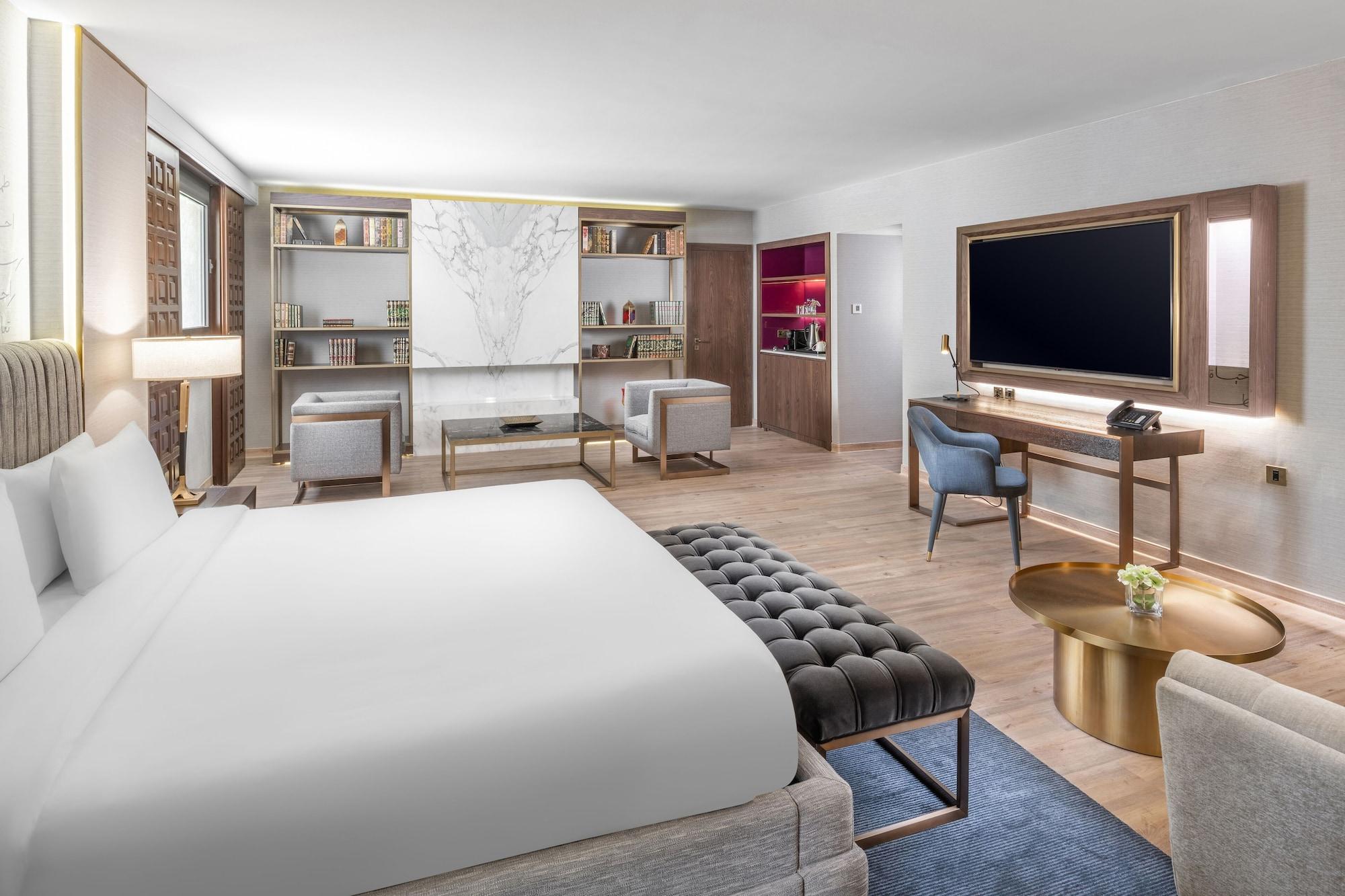 マカレム アイヤッド メッカ ホテル エクステリア 写真 The photo shows a modern hotel room designed with a contemporary aesthetic. The room features a large bed with white bedding, complemented by a stylish tufted bench at the foot of the bed. To the side, there are two comfortable armchairs positioned a