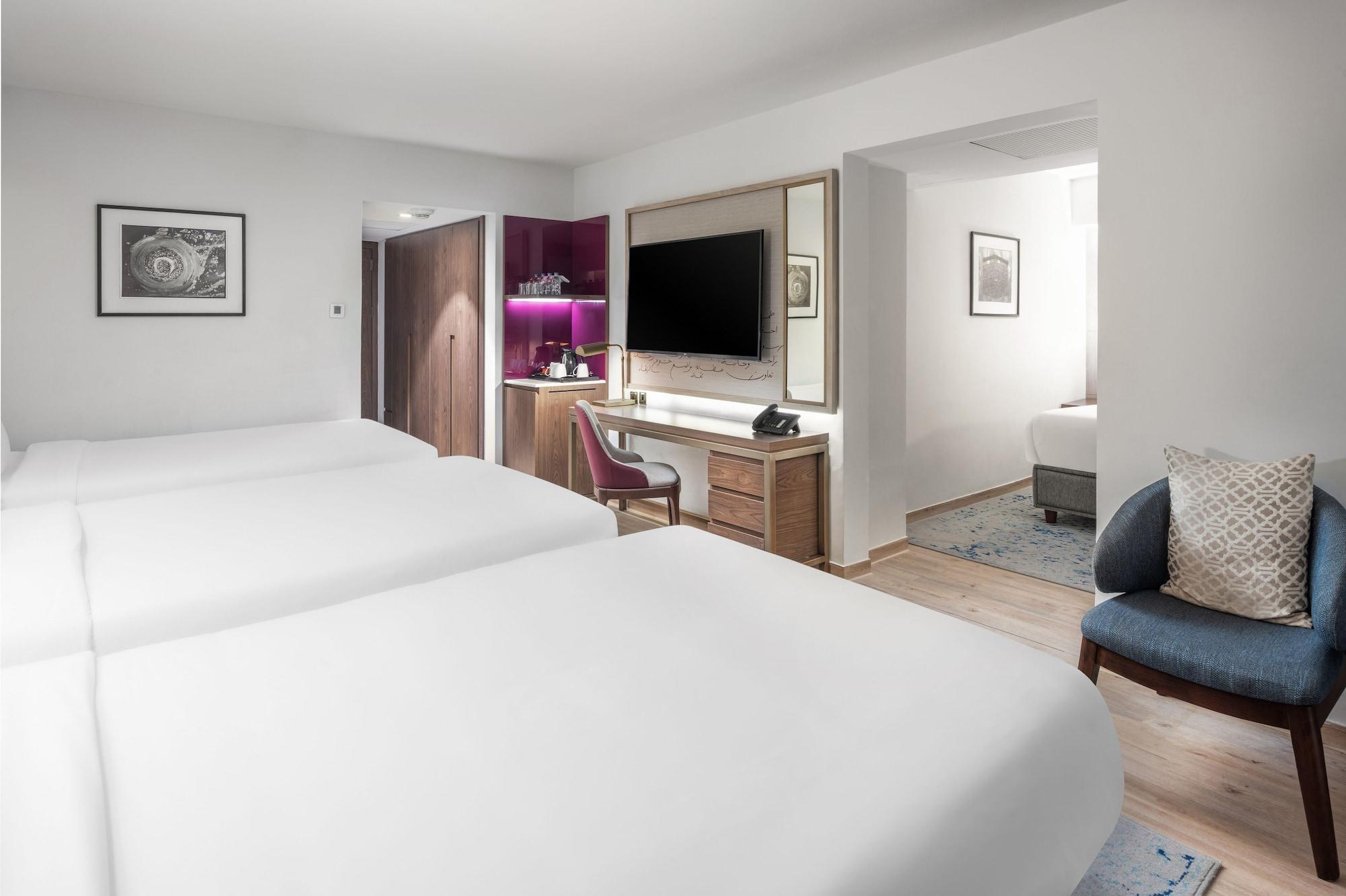 マカレム アイヤッド メッカ ホテル エクステリア 写真 The photo shows a modern hotel room featuring two white beds with smooth linens. The room has a minimalistic design with light-colored walls and wooden flooring. On one side, there is a desk and chair next to a television mounted on the wall, along w