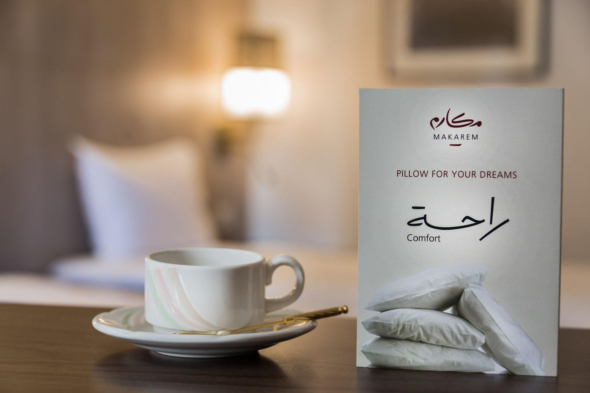 マカレム アイヤッド メッカ ホテル エクステリア 写真 The image features a cozy setting, likely a hotel room. In the foreground, there is a white cup of coffee or tea placed on a small saucer, accompanied by a gold spoon. Beside the cup is a decorative card or sign that reads "Pillow for Your Dreams" an