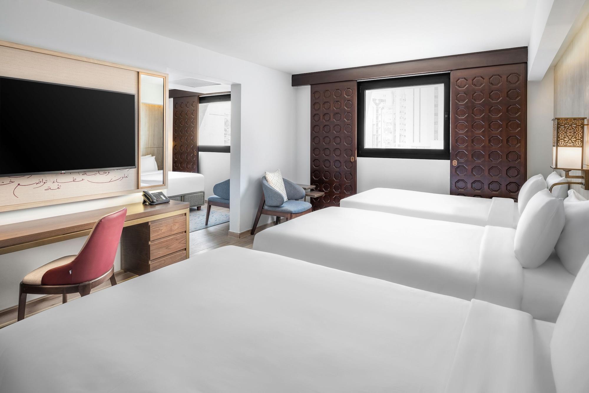 マカレム アイヤッド メッカ ホテル エクステリア 写真 The photo shows a hotel room featuring three beds with white linens. There is a large flat-screen TV mounted on the wall opposite the beds, and a wooden side table beneath it. A red chair is positioned near the TV, while a blue upholstered chair is p