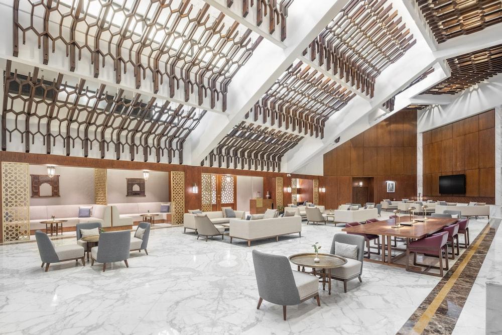 マカレム アイヤッド メッカ ホテル エクステリア 写真 The photo shows a spacious and elegantly designed lobby area, likely within a hotel. The interior features high ceilings with a unique architectural design, showcasing wooden elements arranged in a geometric pattern. The flooring is a polished marble