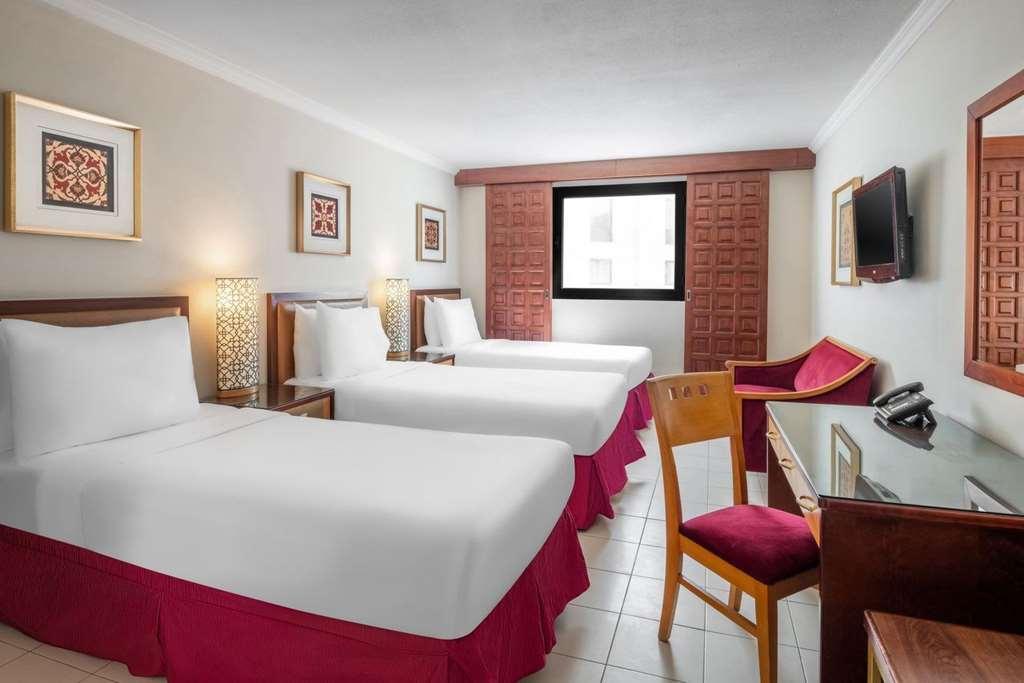 マカレム アイヤッド メッカ ホテル 部屋 写真 The photo shows a hotel room featuring two twin beds with white linens and red bed skirts. There are decorative elements on the walls, including framed artwork. A large window provides natural light, while a desk and a chair are positioned next to it
