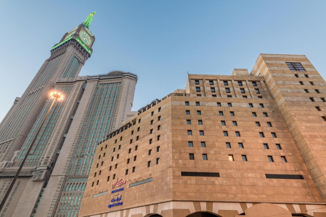 マカレム アイヤッド メッカ ホテル エクステリア 写真 The photo shows a prominent building with a modern architectural design, likely a hotel or commercial establishment, at the forefront. It features a light-colored stone facade with large windows. Behind it, there is a tall skyscraper, which has a dis