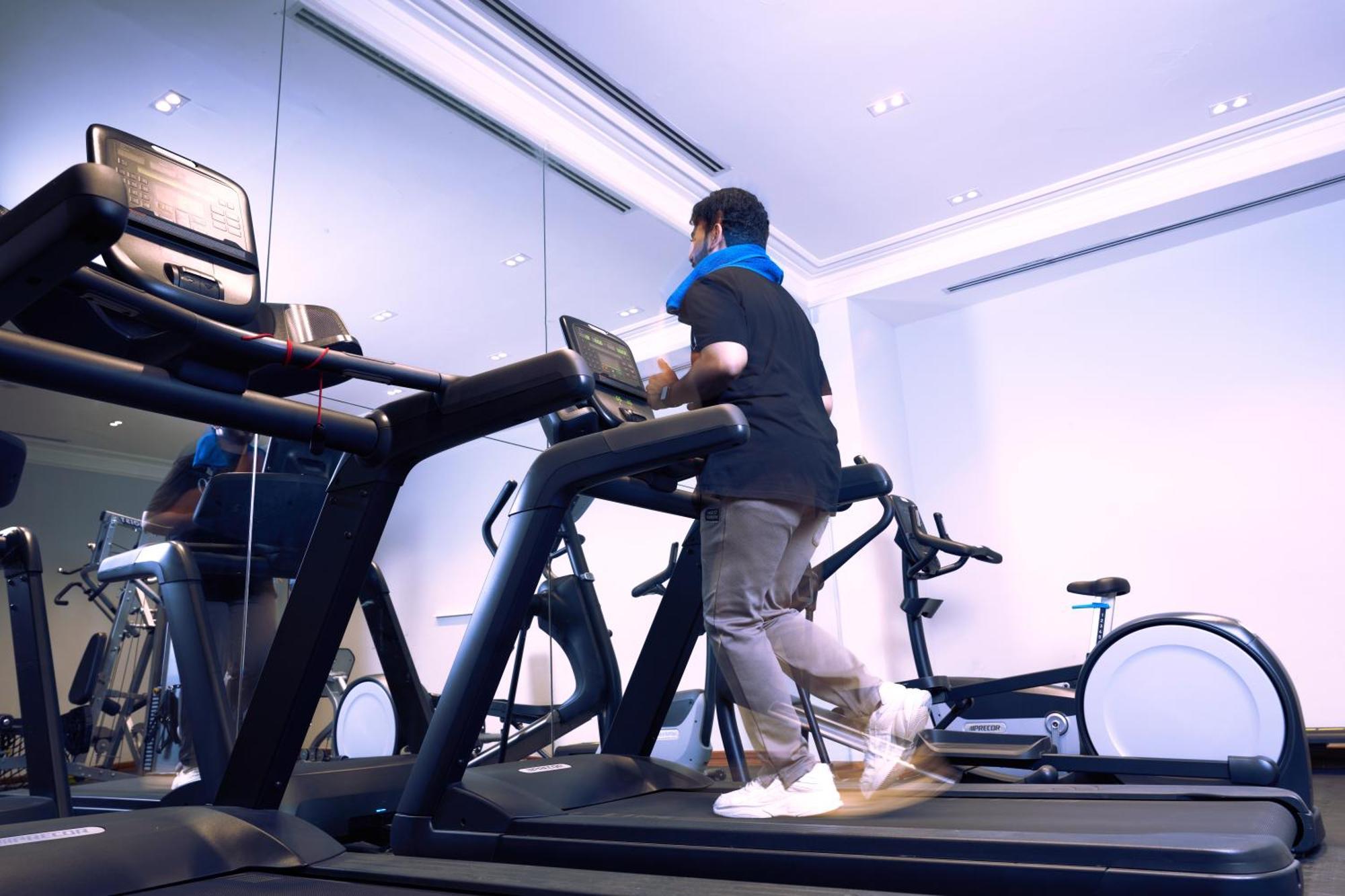 マカレム アイヤッド メッカ ホテル エクステリア 写真 The photo shows a person exercising on a treadmill in a modern gym or fitness room. The individual is wearing a blue neck support while jogging on the treadmill, which is set against a backdrop of large mirrors. There are also additional exercise equ