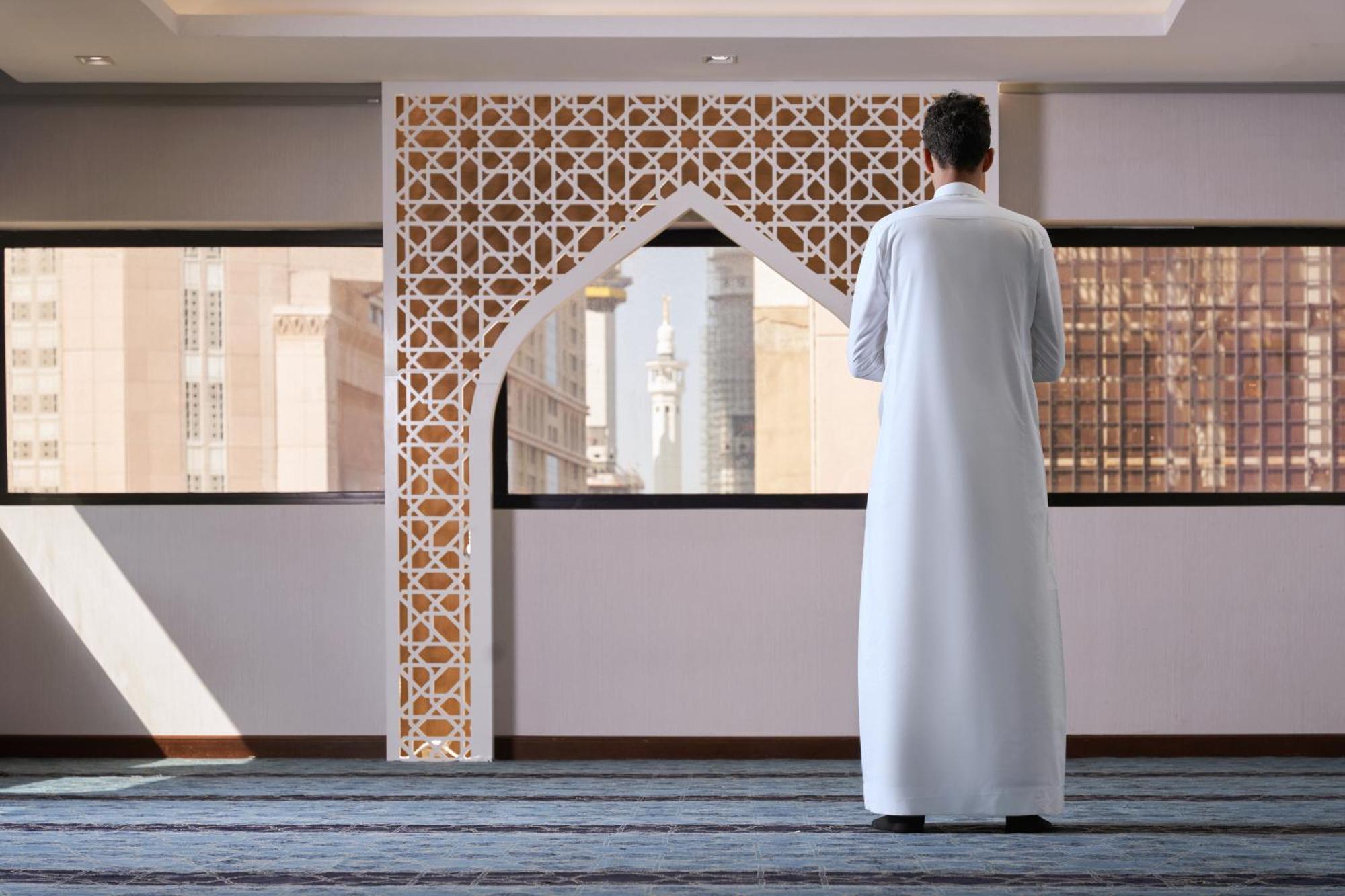 マカレム アイヤッド メッカ ホテル エクステリア 写真 The photo depicts a person standing with their back facing the viewer, wearing a traditional white garment. They are in a spacious interior setting that features an ornate archway with intricate patterns, typical of Islamic architecture. Through the 