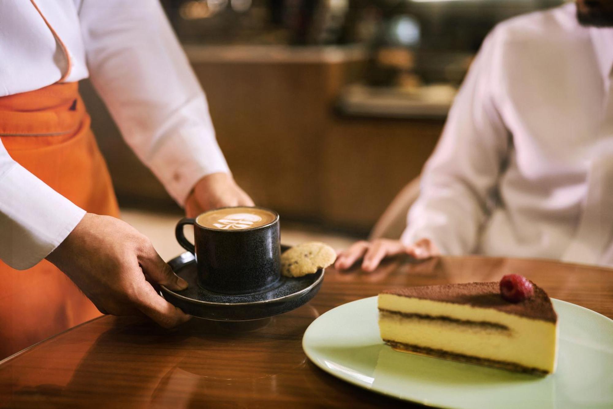 マカレム アイヤッド メッカ ホテル エクステリア 写真 The photo shows a hand presenting a cup of coffee on a saucer, accompanied by a cookie. In the foreground, there is a slice of cake on a light green plate, garnished with a raspberry. The setting appears to be a café or restaurant, with a blurred bac