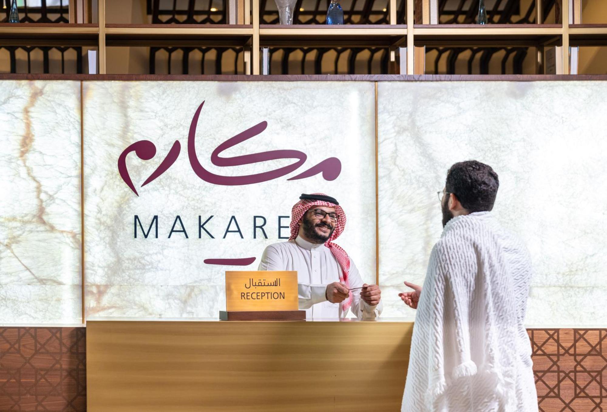 マカレム アイヤッド メッカ ホテル エクステリア 写真 The photo depicts a reception area in a hotel or establishment named "Makarem." In the image, a receptionist, dressed in traditional attire, is smiling and interacting with a guest who is wearing a white robe, commonly known as a thobe or ihram. The 