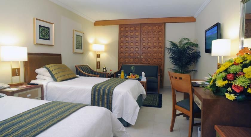 マカレム アイヤッド メッカ ホテル エクステリア 写真 The photo shows a hotel room featuring two single beds with white linens and green and blue striped throw blankets. There is a wooden headboard behind the beds and a decorative wooden partition or wall in the background. A cozy seating area is visibl