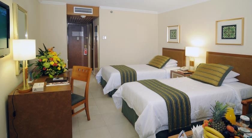 マカレム アイヤッド メッカ ホテル エクステリア 写真 The photo shows an interior of a hotel room featuring two twin beds with white linens and green decorative accents. The walls are painted in a light beige color, and there is a small table next to one of the beds. A desk with a chair is positioned ne