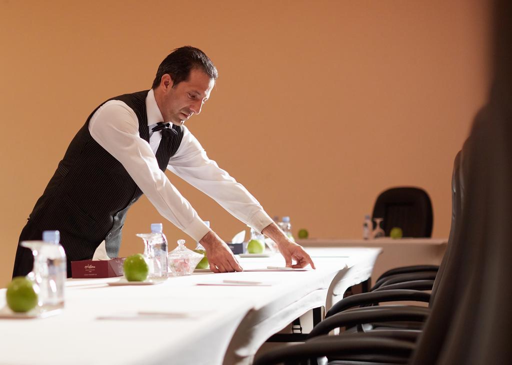 マカレム アイヤッド メッカ ホテル エクステリア 写真 The image shows a waiter or service staff member in a formal attire, which includes a vest and bow tie. He is preparing a long table, likely for an event or meeting. The table is set with items such as water bottles and green apples, and there are se