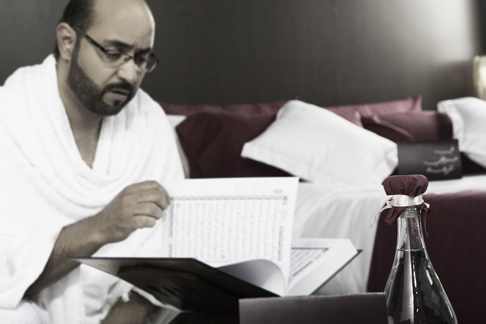 マカレム アイヤッド メッカ ホテル エクステリア 写真 The photo shows a man engaged in reading a book or document, seated in a room with a relaxed atmosphere. He is wearing traditional white attire, often associated with specific cultural or religious practices. In the foreground, there is a bottle, pos